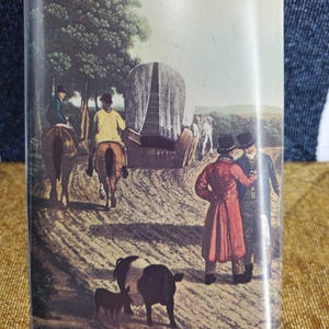 May include: Rectangular tin featuring a vintage illustration of a rural scene with people on horseback, a covered wagon, and figures in coats. The tin has a red border.