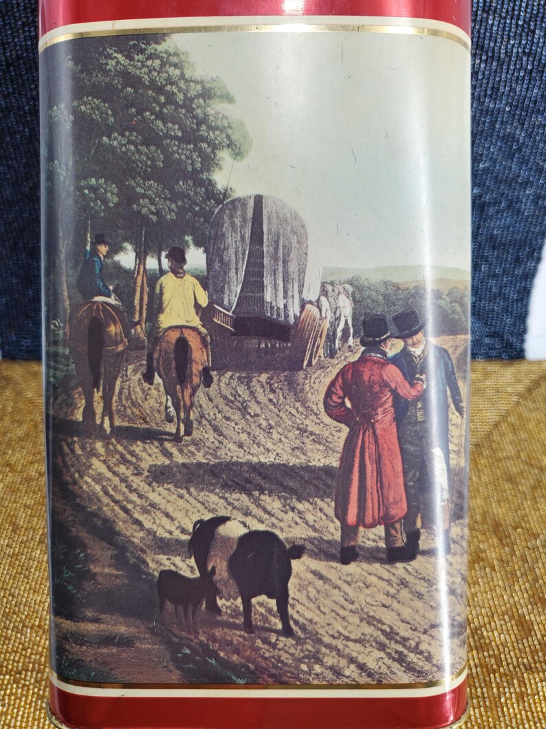 May include: Rectangular tin featuring a vintage illustration of a rural scene with people on horseback, a covered wagon, and figures in coats. The tin has a red border.