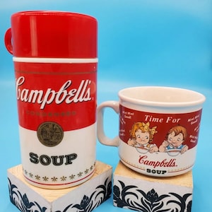 Könnte beinhalten: Zwei rote und weiße Campbell's Suppenbehälter. Einer ist eine Tasse mit dem Text "Time For Campbell's Soup" und einem Cartoon-Bild von zwei Kindern, die Suppe essen. Der andere ist eine rote Thermoskanne mit dem Text "Campbell's Soup".