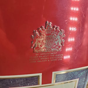 May include: Red metal container with a gold emblem and text. The emblem features a royal crest. Text includes "By Appointment to Her Majesty The Queen" and "John Walker & Sons Ltd."