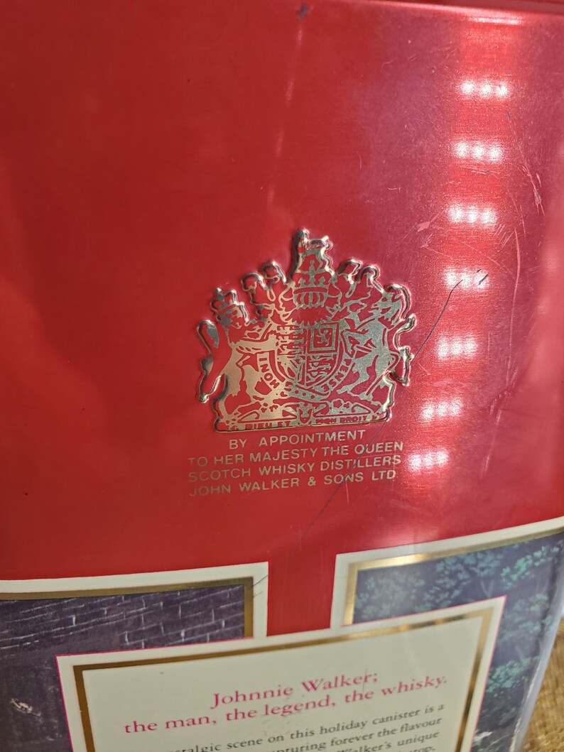 May include: Red metal container with a gold emblem and text. The emblem features a royal crest. Text includes "By Appointment to Her Majesty The Queen" and "John Walker & Sons Ltd."