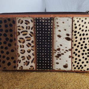 Puede incluir: Un monedero rectangular con cierre de cremallera, con un diseño de patchwork. La parte delantera presenta cinco paneles verticales con estampados de animales y patrones de lunares en marrón, negro y crema. Los bordes están ribeteados en cuero marrón claro.
