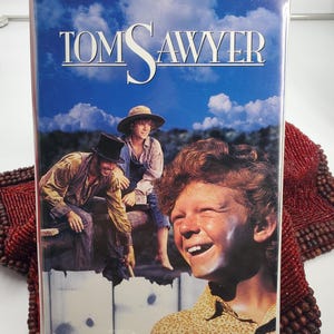 Puede incluir: Una cinta de vídeo VHS de la película "Tom Sawyer" con un fondo de cielo azul. La portada presenta a un niño sonriente y otros personajes. El título está en letras blancas grandes. La cinta está etiquetada como "Family Treasures" por MGM/UA.