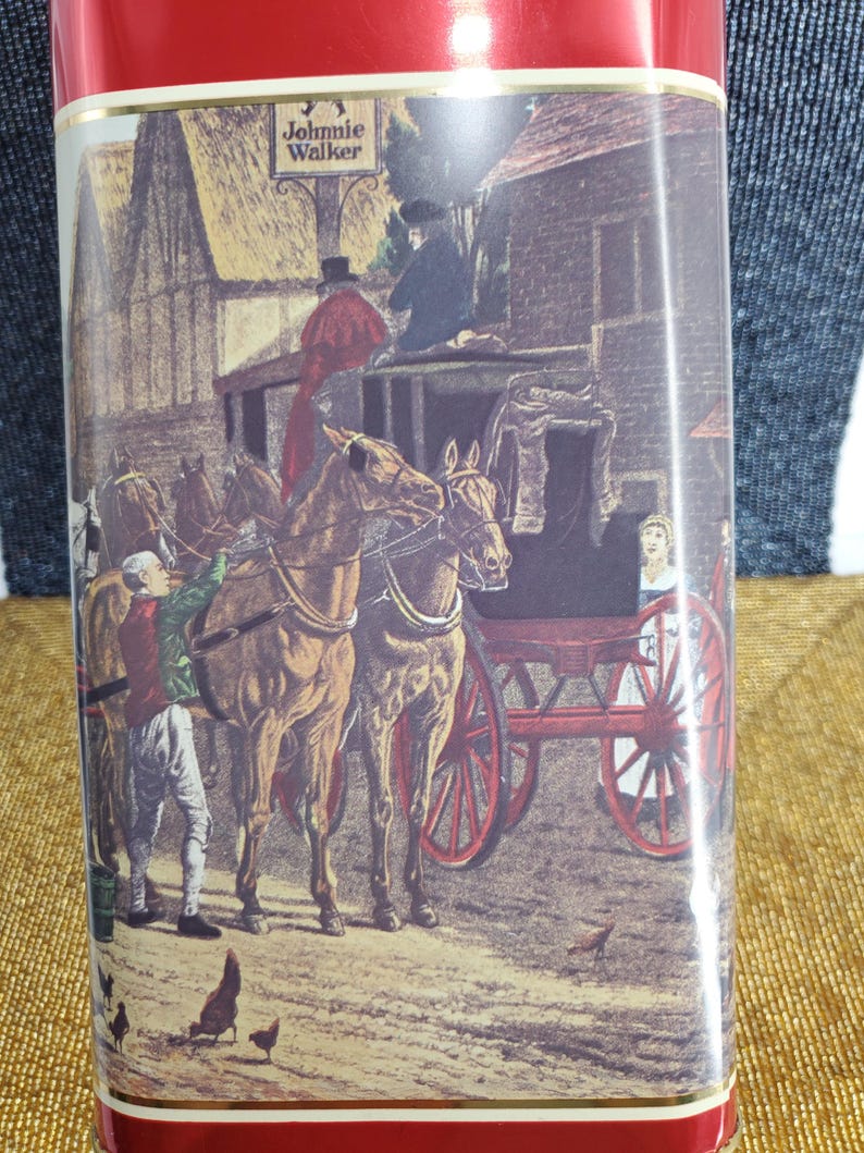 May include: A red container with a detailed illustration of a horse-drawn carriage, people, and buildings. The text "Johnnie Walker" is at the top. The scene is set in a rural environment with chickens.