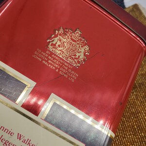 May include: A red rectangular tin with gold trim and text. The tin features a gold emblem and the text "BY APPOINTMENT TO HER MAJESTY THE QUEEN SCOTCH WHISKY DISTILLERS JOHN WALKER & SONS LTD".