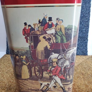 May include: Red metal umbrella stand featuring a vintage illustration of a horse-drawn carriage and people in period clothing. The word "Red" is displayed in gold lettering at the top.