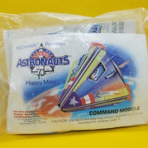 Könnte beinhalten: Eine weiße Plastiktüte mit einem Happy Meal Spielzeug. Das Spielzeug ist ein blaues, grünes und rotes Raumschiff mit einem weißen Stern an der Seite. Auf der Tüte steht "McDonald's Presents Young Astronauts" und "Happy Meal". Die Tüte enthält auch einen Warnhinweis, dass das Spielzeug kleine Teile enthalten kann und nicht für Kinder unter 3 Jahren geeignet ist.