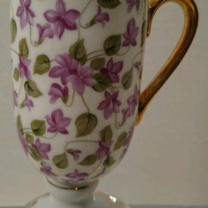 May include: A white teacup with a gold handle and base, adorned with a floral pattern of purple flowers and green leaves. The cup sits on a clear, decorative base, creating an elegant aesthetic.