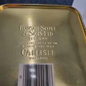 May include: A gold-coloured tin box with embossed text. The text reads "HUDSON-SCOTT & SONS-LTD (EST. 1799)", "MANUFACTURERS OF TIN BOXES SINCE 1866", and "CARLISLE ENGLAND". A barcode label is affixed to the bottom.