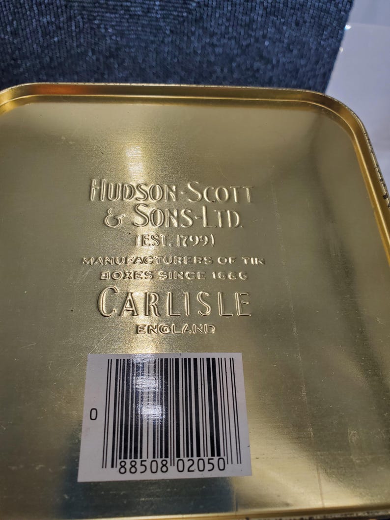 May include: A gold-coloured tin box with embossed text. The text reads "HUDSON-SCOTT & SONS-LTD (EST. 1799)", "MANUFACTURERS OF TIN BOXES SINCE 1866", and "CARLISLE ENGLAND". A barcode label is affixed to the bottom.