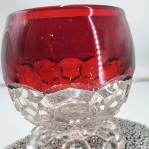 May include: A ruby red glass bowl with a clear, faceted base. The bowl's upper portion is a deep red, while the base is clear with a geometric pattern. The bowl sits on a textured surface.