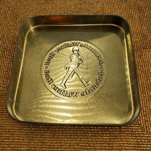 May include: A gold-coloured metal tray with rounded corners. The centre features a circular embossed design of a man walking, surrounded by text. The tray has a slightly worn appearance.