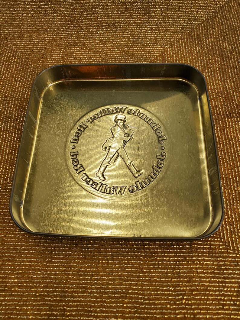 May include: A gold-coloured metal tray with rounded corners. The centre features a circular embossed design of a man walking, surrounded by text. The tray has a slightly worn appearance.
