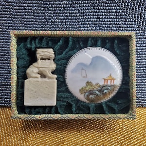 Puede incluir: Una caja decorativa con un sello de piedra tallada y una pieza de cerámica redonda pintada. El sello presenta una figura de león. La pieza de cerámica representa un paisaje con montañas, un barco y una pagoda.
