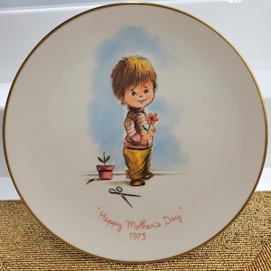 Op de afbeelding: Een rond wit decoratief keramieken bord met een goudkleurige rand. Het midden toont een kleurrijke illustratie van een kind dat een oranje bloem vasthoudt, met een kleine potplant en een schaar erbij. De tekst luidt "Happy Mother's Day 1973".