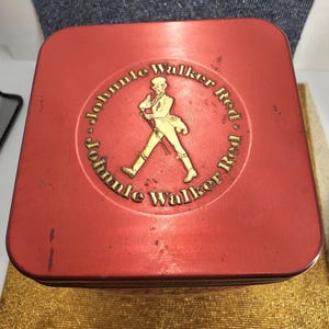May include: A red rectangular tin featuring the words "Johnnie Walker Red" in gold lettering and a gold figure of a man walking. The tin has a slightly worn appearance.