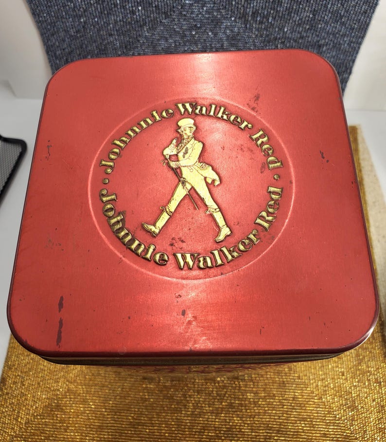 May include: A red rectangular tin featuring the words "Johnnie Walker Red" in gold lettering and a gold figure of a man walking. The tin has a slightly worn appearance.