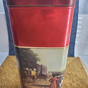 May include: A tall, rectangular red tin with a matching lid. The lower half of the tin features a pastoral scene with figures, horses, and a covered wagon. The tin is likely for storage or display.