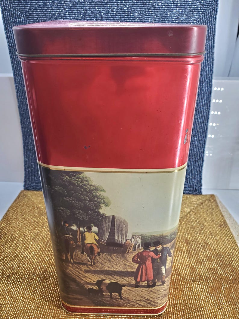 May include: A tall, rectangular red tin with a matching lid. The lower half of the tin features a pastoral scene with figures, horses, and a covered wagon. The tin is likely for storage or display.