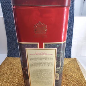 May include: A red rectangular tin with a gold emblem and text. The tin features a nostalgic scene with text that reads "Johnnie Walker: the man, the legend, the whisky." The tin is likely for storing whisky.