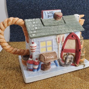 May include: A ceramic teapot shaped like a fishing shack, featuring a red boat, anchor, and the words "LIVE BAIT." The handle is a rope design, and the roof is green. The teapot has a nautical theme.
