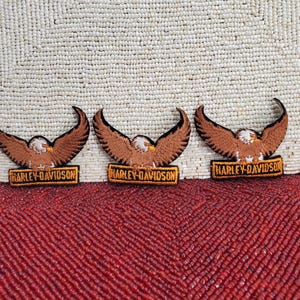 May include: Three identical embroidered patches featuring a brown eagle with outstretched wings positioned above a rectangular orange and black banner. Each patch displays the text HARLEY-DAVIDSON in black capital letters on the banner.