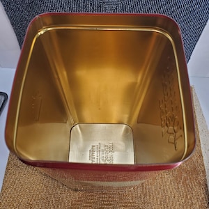 May include: A rectangular tin with a red exterior and a gold interior. The tin is empty and has a slightly trapezoidal shape. The interior is a shiny gold colour, and the red exterior has a glossy finish. The bottom of the tin has text.