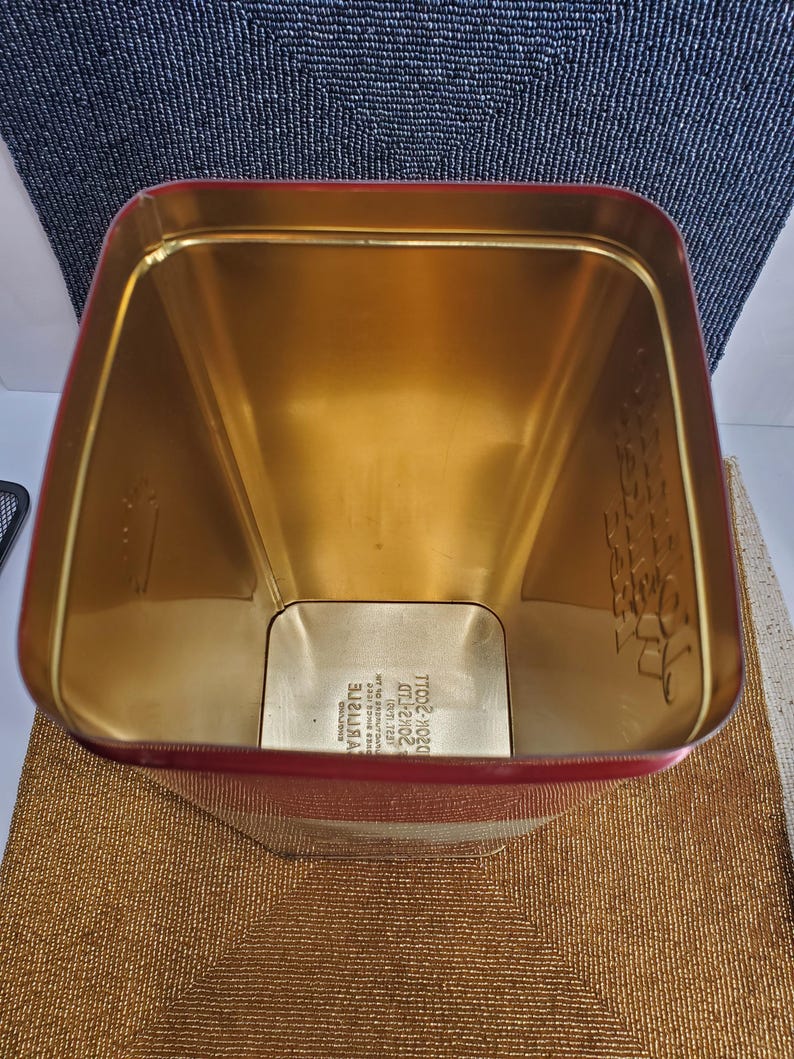 May include: A rectangular tin with a red exterior and a gold interior. The tin is empty and has a slightly trapezoidal shape. The interior is a shiny gold colour, and the red exterior has a glossy finish. The bottom of the tin has text.