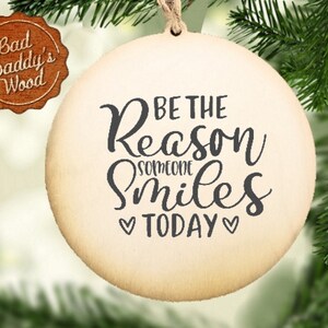 May include: A round wooden ornament with the text "Be the reason someone smiles today" in black script lettering. The ornament is hanging on a Christmas tree.