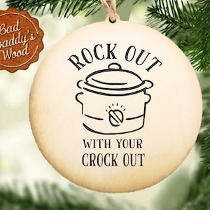 Peut inclure: Une décoration ronde en bois avec le texte "Rock Out With Your Crock Out" et un dessin d'une mijoteuse.
