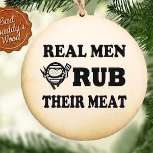 Può includere: Ornamento natalizio rotondo in legno chiaro con la scritta nera "REAL MEN RUB THEIR MEAT". Al centro c'è una grafica di un barbecue con utensili. Ha un cordino di iuta per appenderlo e il logo "Bad Daddy's Wood".