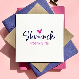 May include: A white square card with the text "Shmuncki Poem Gifts" in a handwritten style, with a pink heart above the word "Shmuncki". The card is on top of a stack of notebooks and envelopes in shades of pink, blue, and tan.