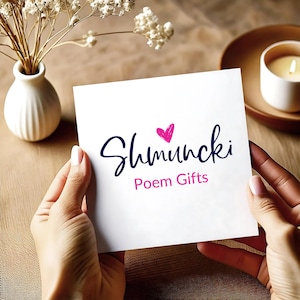 May include: A white card with the text "Shmuncki Poem Gifts" in a handwritten style, featuring a pink heart. The card is held by two hands, with a blurred background including a vase with flowers, a candle, and a wooden surface.