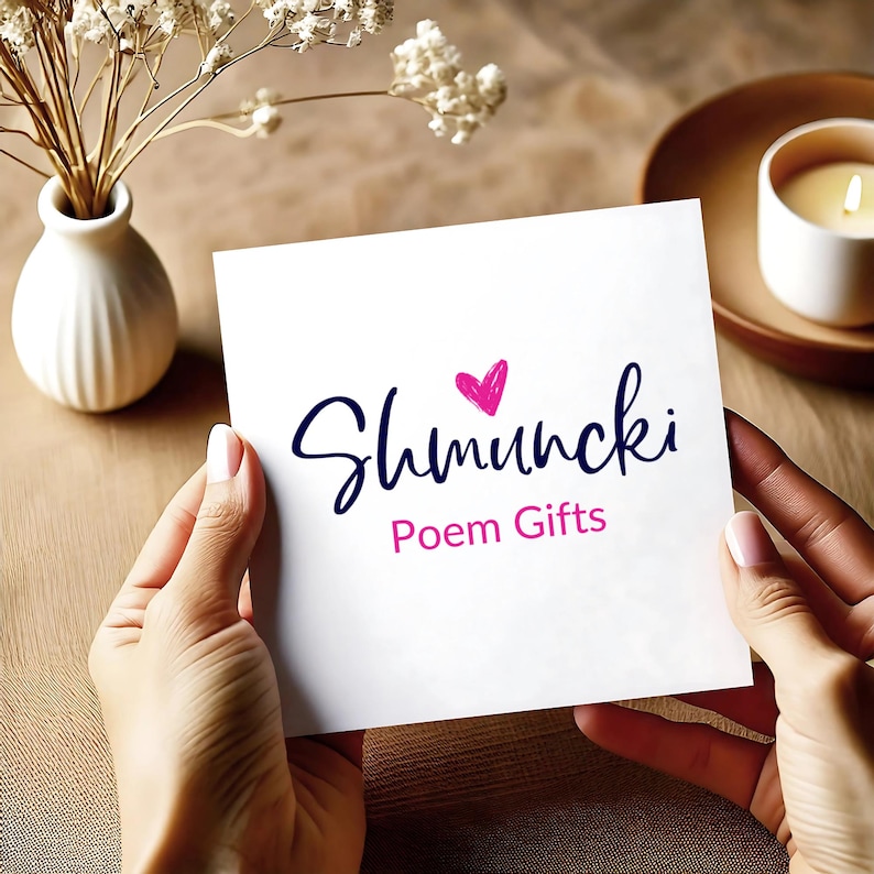 May include: A white card with the text "Shmuncki Poem Gifts" in a handwritten style, featuring a pink heart. The card is held by two hands, with a blurred background including a vase with flowers, a candle, and a wooden surface.