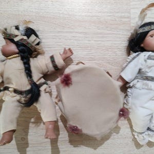 Könnte beinhalten: Zwei Indianerpuppen mit schwarzen Zöpfen und traditioneller Kleidung. Eine Puppe trägt ein cremefarbenes Outfit mit Federkopfschmuck und hält einen Stock. Die andere Puppe trägt ein weißes Outfit mit Federkopfschmuck. Zwischen ihnen befindet sich eine beige Trommel mit roten Akzenten.