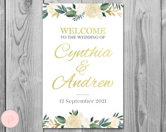 May include: White wedding welcome sign with gold lettering. The sign says "Welcome to the wedding of Cynthia & Andrew 12 September 2021". The sign is decorated with white and green floral designs.
