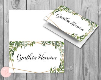 May include: A white place card with a gold geometric frame and green watercolor foliage. The name "Cynthia Honma" is written in black cursive lettering.