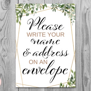 May include: White sign with gold geometric frame and green foliage. The sign says "Please write your name & address on an envelope".