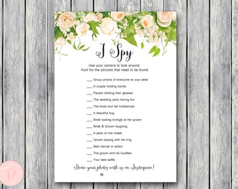 May include: A white card with a floral border and the text "I Spy" printed on it. The card lists a series of things to find at a wedding reception, such as a couple holding hands, people clinking glasses, and the bride and groom laughing. The card also includes the text "Share your photos with us on Instagram! #"