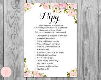 May include: A white card with pink floral borders and the text "I Spy" printed in black. The card lists a series of wedding-themed photo scavenger hunt prompts, such as "Group photos of everyone at your table" and "The groom and his buddies".