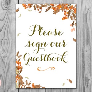 May include: A white sign with brown and orange fall leaves and berries around the border. The text on the sign reads "Please sign our Guestbook".