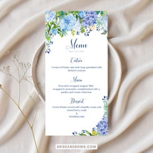 May include: A white menu card with a floral design featuring blue hydrangeas. The menu includes the words "Menu", "Entree", "Main", and "Dessert", with a list of dishes. The bottom of the card has the text "BRIDEANDBOWS.COM".