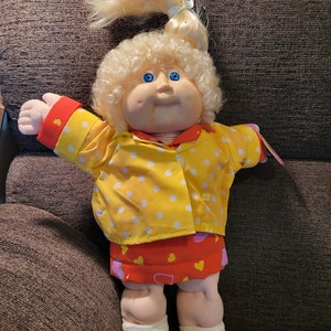 May include: A Cabbage Patch Kid doll with blonde curly hair, wearing a yellow polka dot shirt, a red skirt with pink hearts, and white shoes.