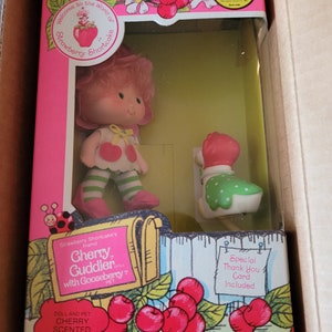 May include: Strawberry Shortcake's friend, Cherry Guddler doll with Gooseberry pet. The doll is wearing a pink dress with green and white stripes, a yellow bow, and white shoes. The doll is holding a red and white comb. The doll is packaged in a pink and green box with a picture of a mailbox and a goose. The box also includes a special thank you card.