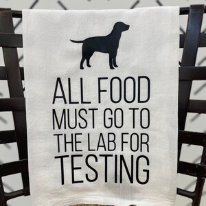 May include: White kitchen towel with a black silhouette of a dog and the text "ALL FOOD MUST GO TO THE LAB FOR TESTING". The towel is draped over a chair with a woven seat.