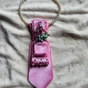 May include: A pink satin tie with a decorative brooch featuring a silver figure and green and clear accents. The tie is adorned with a small pocket and embellished with pearls and green beads. A pearl necklace is attached to the tie.