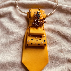 May include: A mustard yellow tie with a pearl necklace. The tie features a jeweled peacock brooch and small pearls and blue gems. The necklace is a strand of small, white pearls.