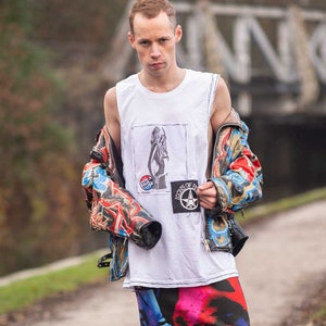 Puede incluir: Una persona lleva una camiseta blanca sin mangas con un estampado gráfico en blanco y negro, una chaqueta con estampado colorido, una falda estampada en negro y rosa, calcetines a rayas blanco y negro y botas negras. La camiseta tiene un parche negro con una estrella y el texto "Icons of the 80s". La falda tiene el texto "That's not gender, that's a style".