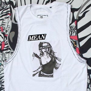 Può includere: Canotta bianca con grafica in bianco e nero di una donna che tiene un'arma. La parola "MEAN" è sopra la grafica e l'hashtag "#BADGIRL" è sul volto della donna.