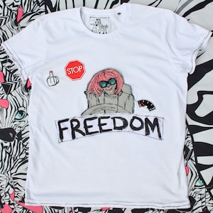 Può includere: Maglietta bianca con un cartello di stop rosso, un dito medio, una donna che guida un'auto e la parola "FREEDOM" scritta in nero.